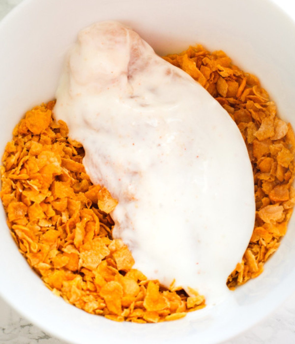 Marinated chicken breast coated in crushed cornflakes in a white bowl.