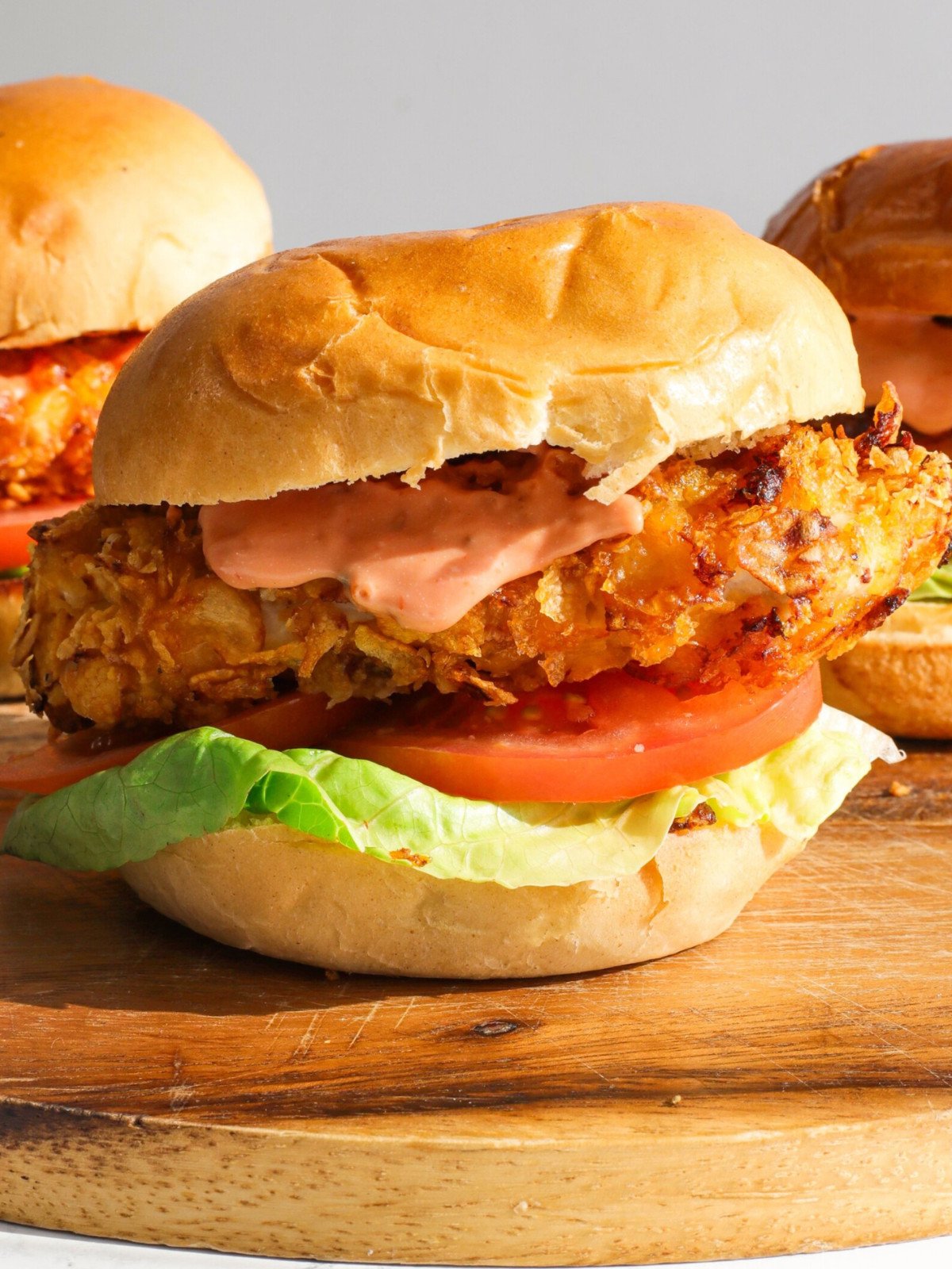 Three crispy chicken burgers filled with tomatoes, lettuce and burger sauce on a wooden board.
