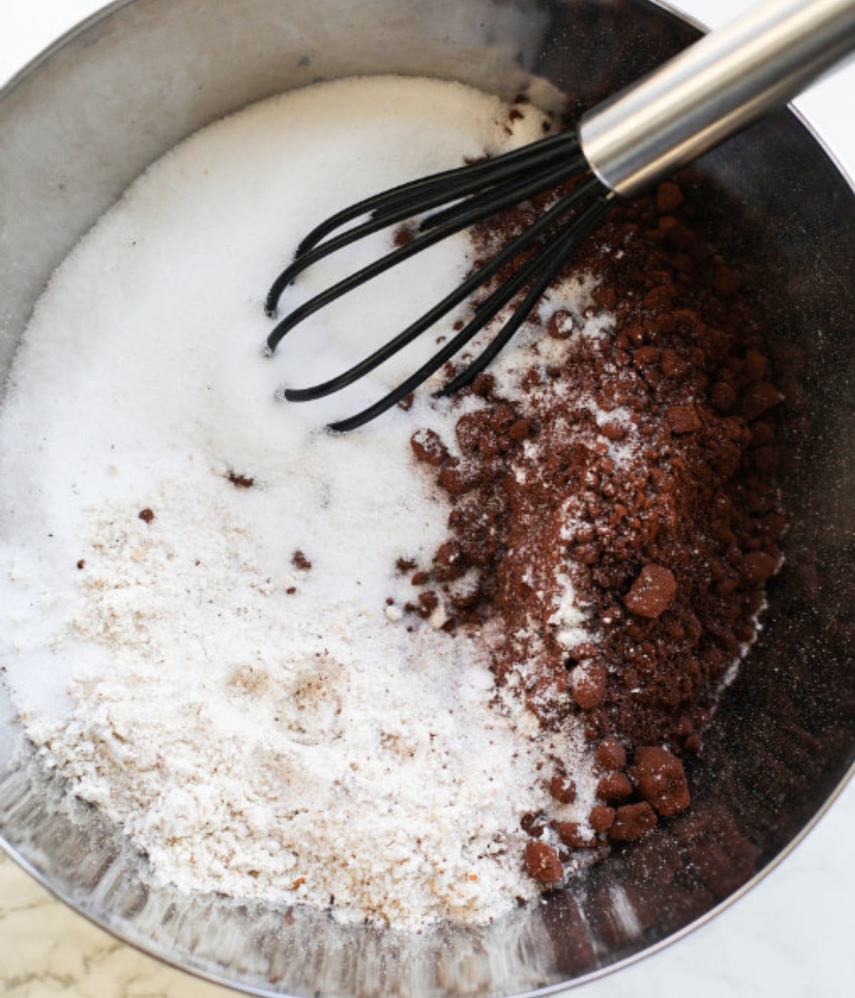 Flour, cocoa powder, and sugar in a silver bowl with a mini black whisk.