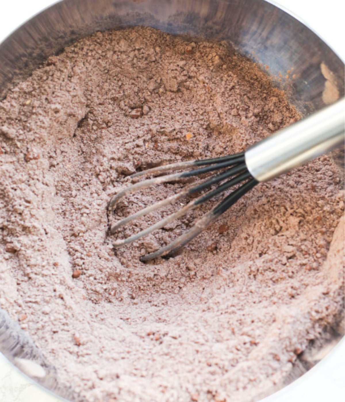 Mixed flour, cocoa powder, and sugar in a silver bowl with a mini black whisk.