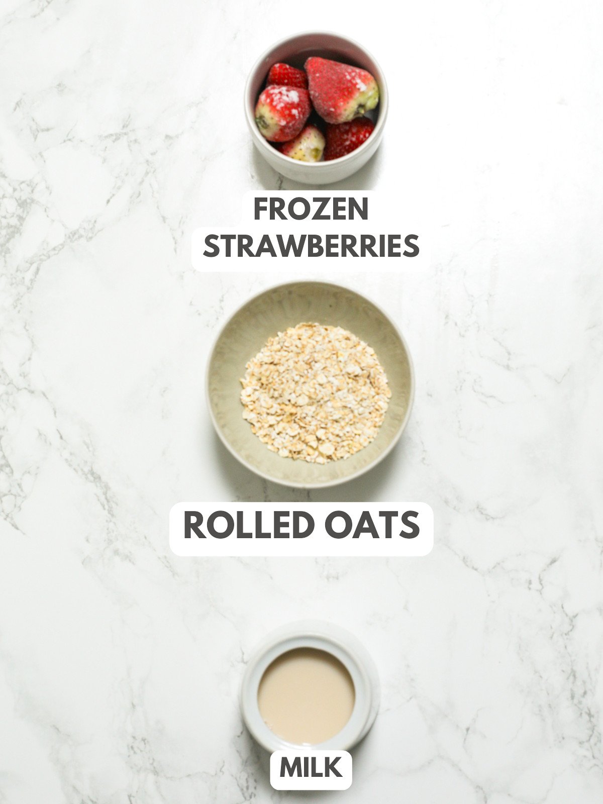 Frozen strawberries, oats and milk laid out on a grey marble table.
