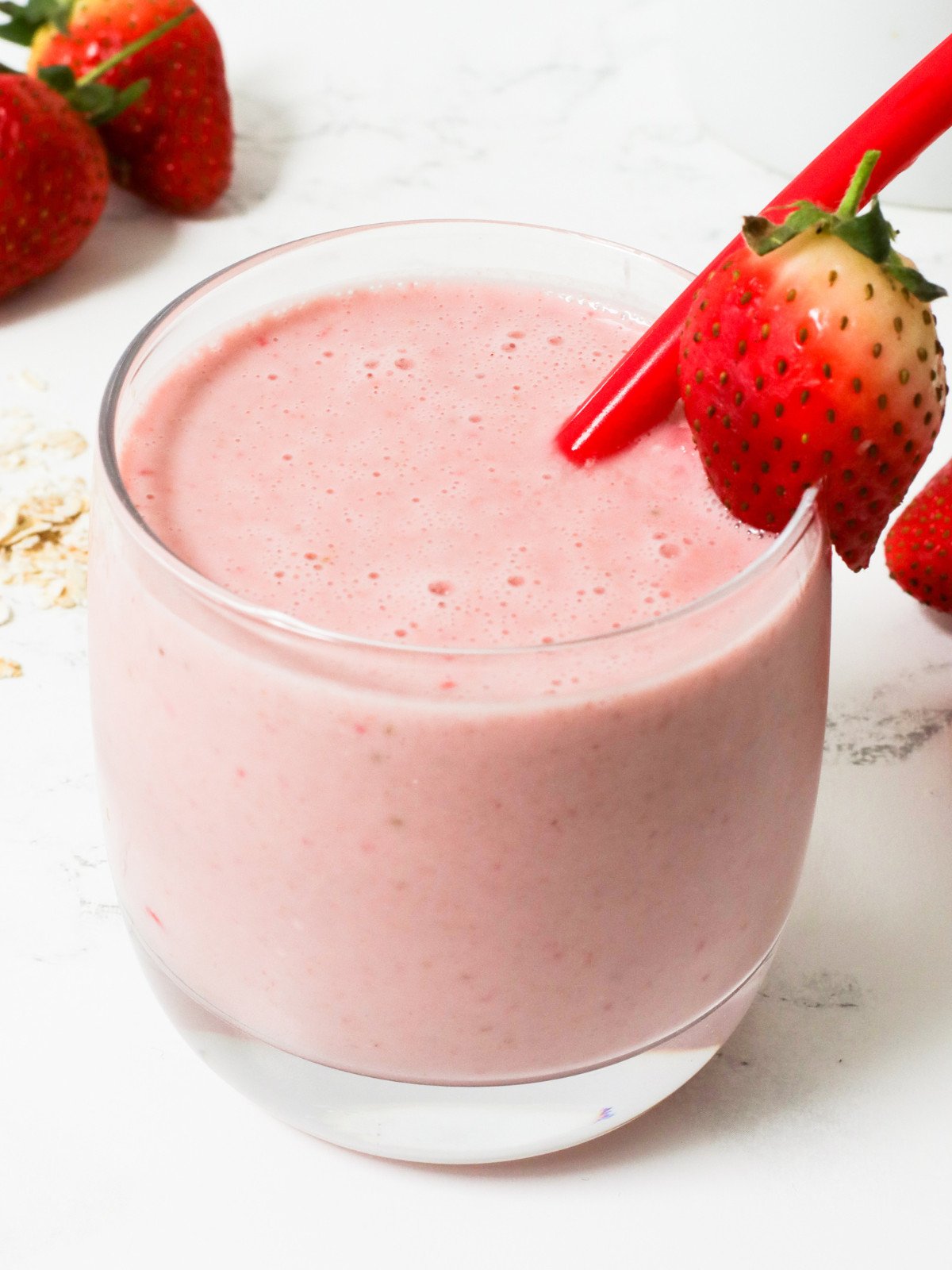 Strawberry oat smoothie in a glass cup with a strawberry and red straw.
