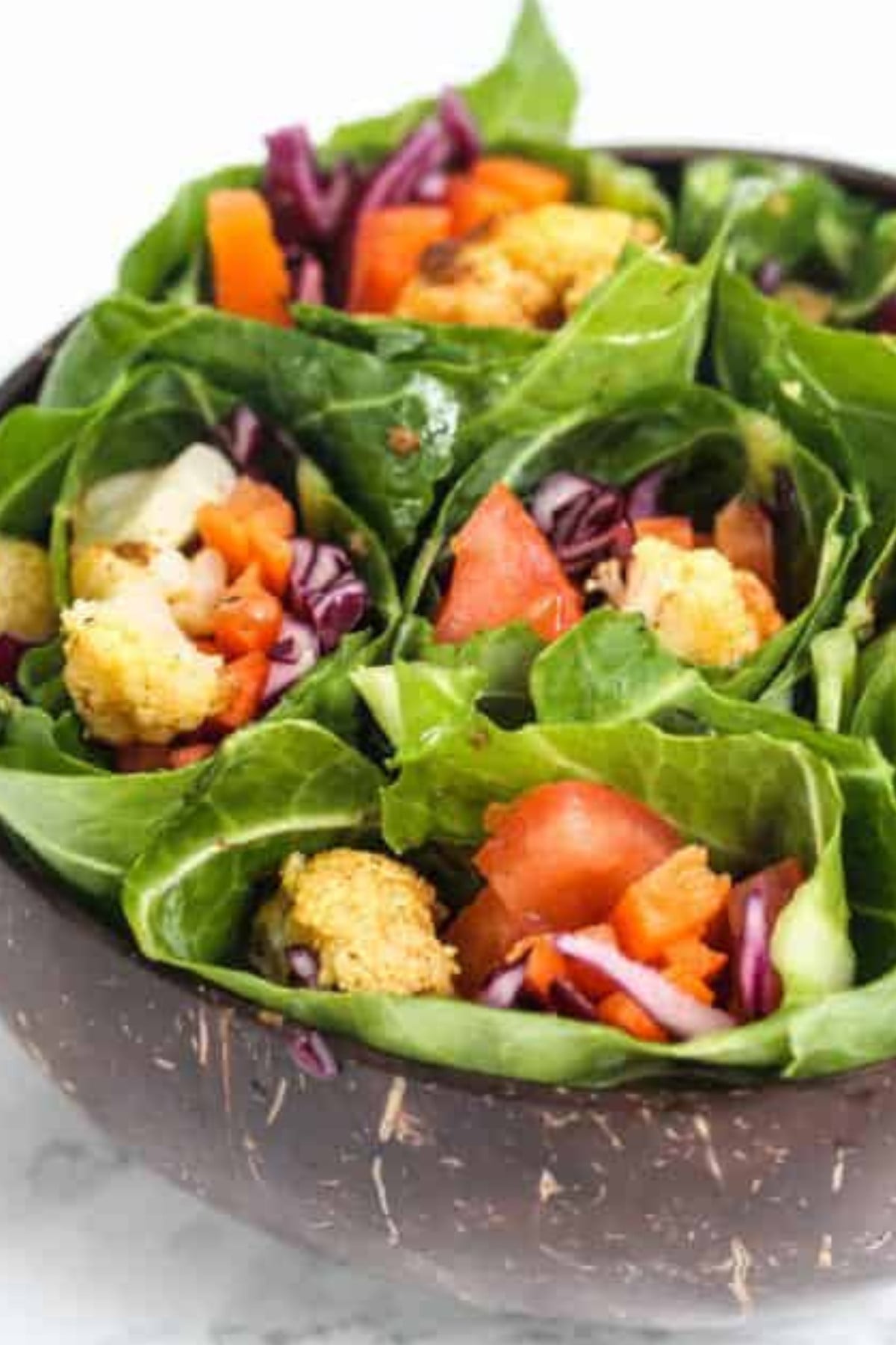 Cauliflower florets wrapped in collard greens with tomatoes, red cabbage and carrots in a brown bowl.
