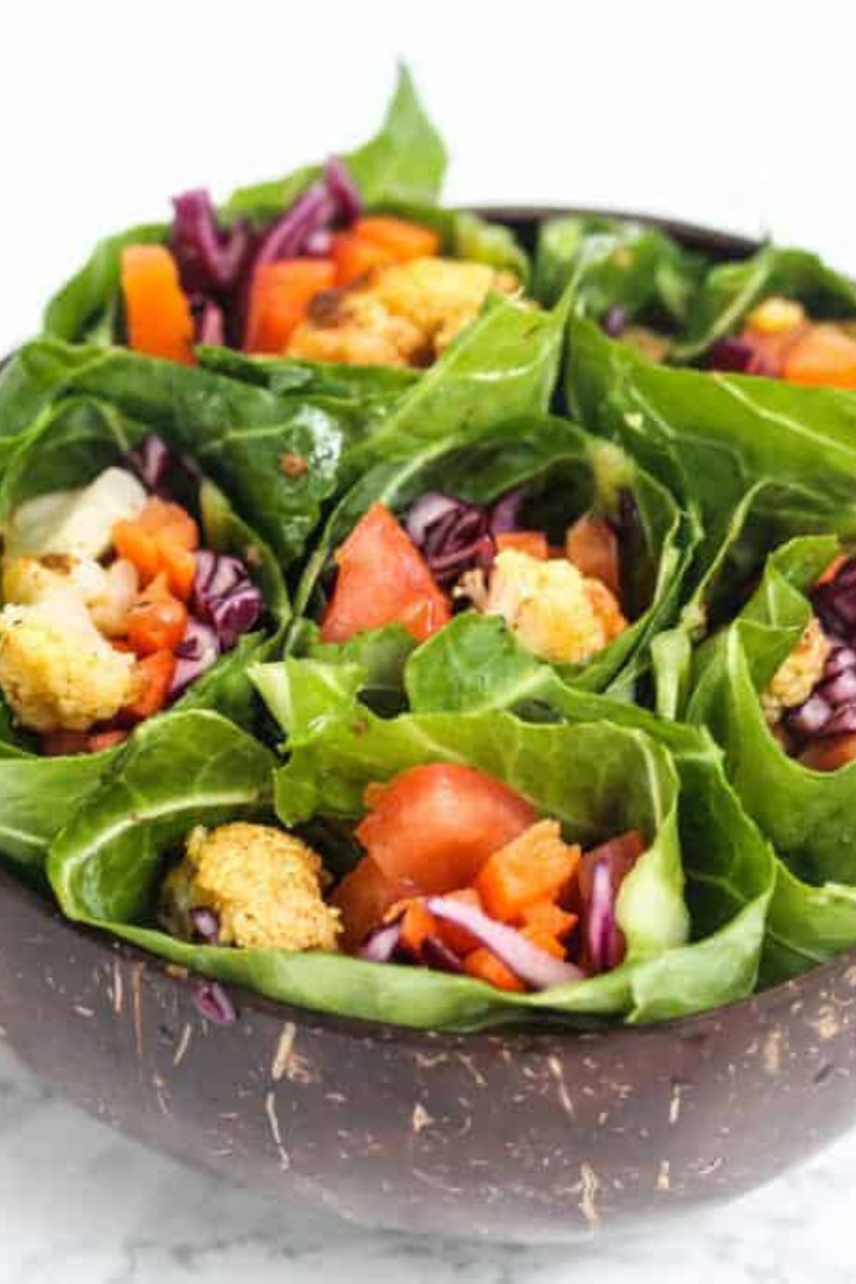 Cauliflower wrapped in collard greens with tomatoes, red cabbage and carrots in a brown bowl.