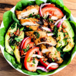 Chicken slices, tomato, avocado, red onions and lettuce in a white bowl.