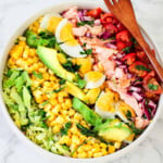 Salmon, avocado, corn, lettuce, boiled eggs, red onions and tomatoes in a white bowl. A wooden fork on top of the plate.
