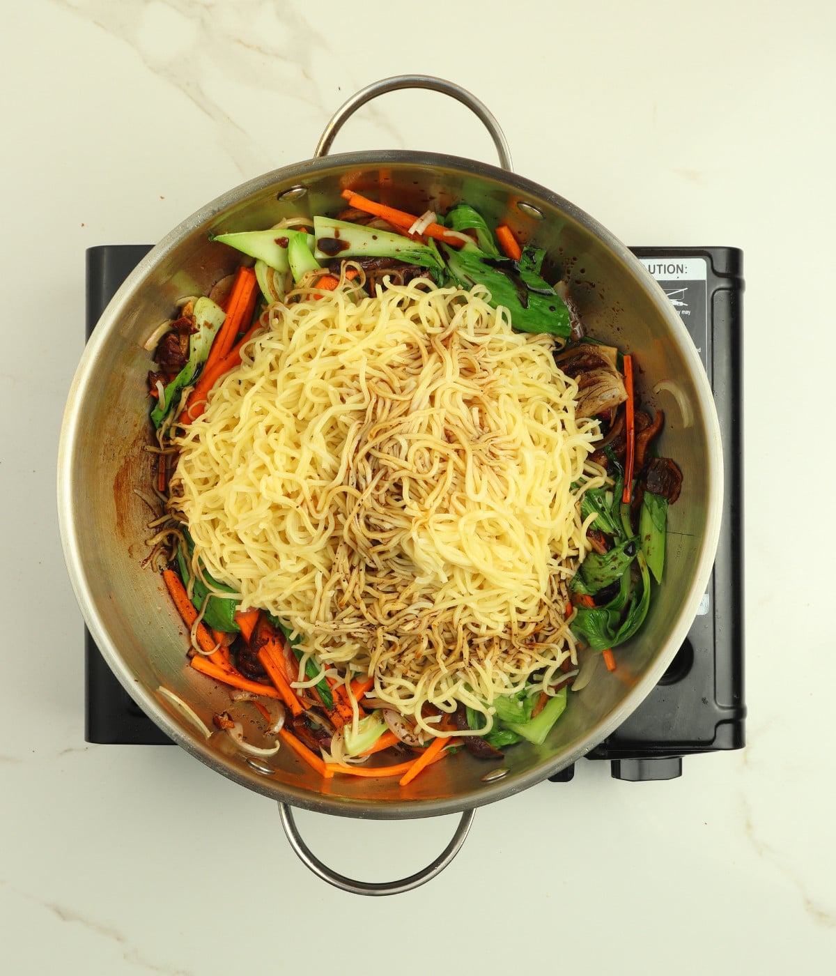 Mushroom, carrots, chow mein, pak choi, soy sauce and oil in a silver wok.