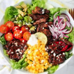 Steak, tomato, sweetcorn, red onions, lettuce and avocado in a white bowl.