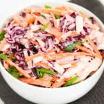 Chopped white cabbage, red cabbage, carrots, onions and green onions in a white bowl on a grey cloth.