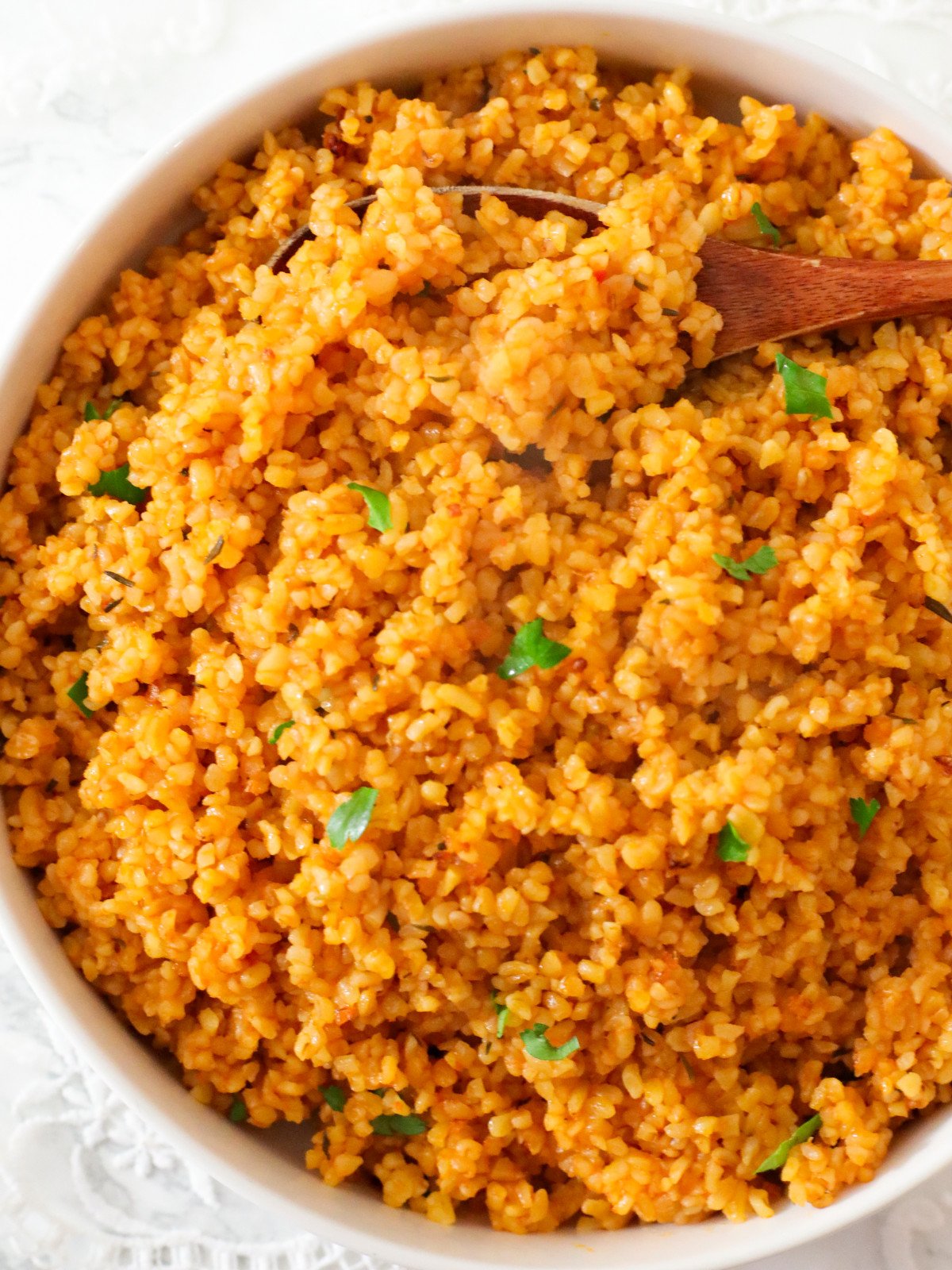 Bulgur jollof in a white bowl with fresh herbs and a brown spoon.