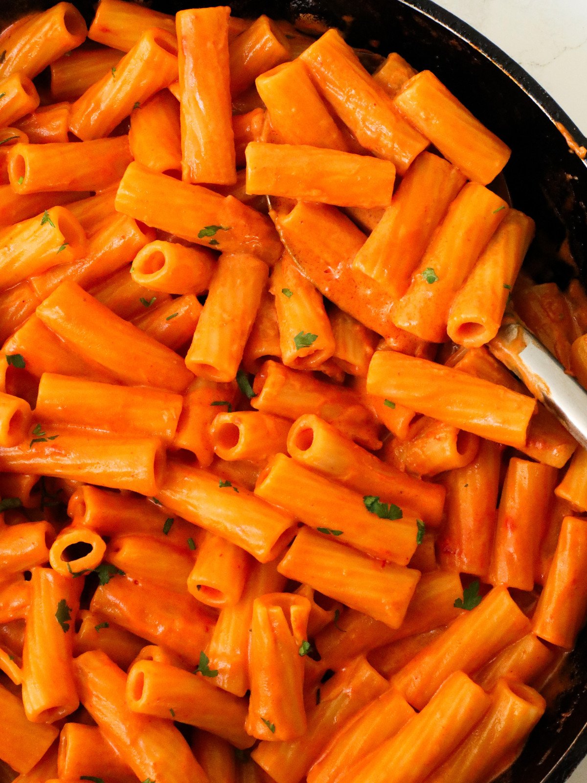Creamy tomato rigatoni and herbs in a black pan with a silver serving spoon.