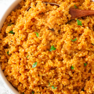 Jollof bulgur wheat served in a white bowl with herbs and a wooden spoon.