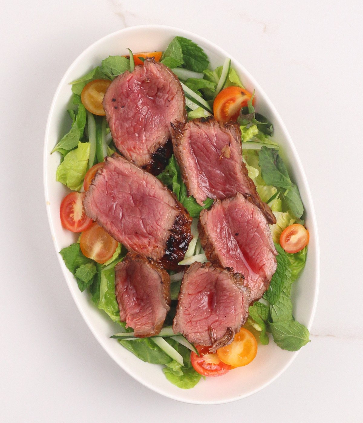 Lettuce, cucumber, tomatoes and sirloin steak slices in a white oval bowl.