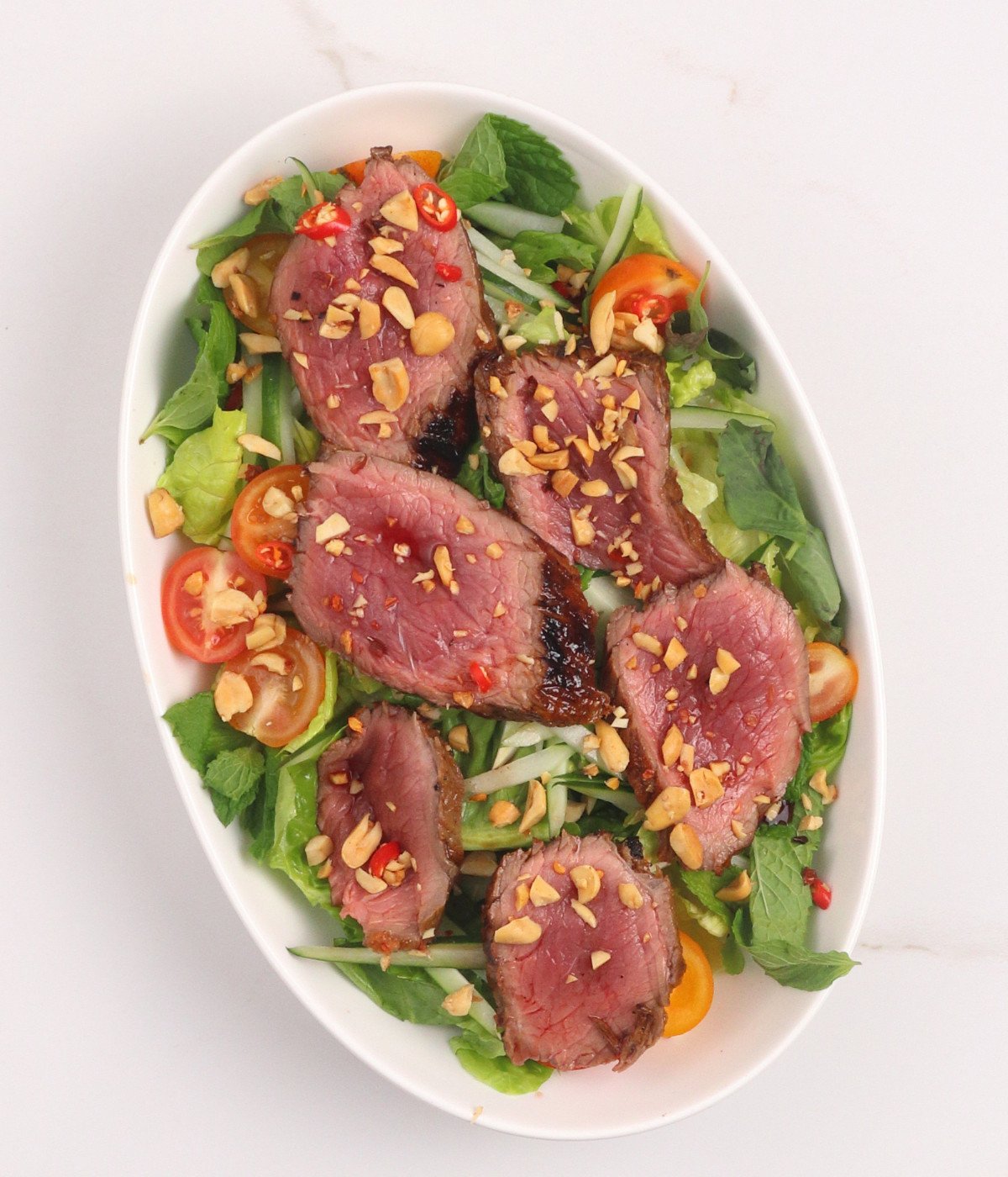 Lettuce, cucumber, tomatoes, peanuts and sirloin steak slices in a white oval bowl.