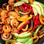 Shrimp, capsicum peppers, avocado slices and lime wedges in a black pan.