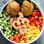 Shrimp, hasselback potatoes, cucumber, tomatoes and corn in a white bowl.
