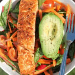 Salmon, avocado, carrots, tomatoes and leaves in a white bowl.