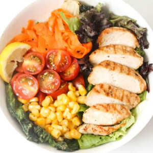 Chicken, tomatoes, sweetcorn, lettuce, carrots and lemon in a white bowl.