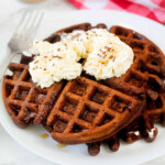 Chocolate waffle topped with whipped cream and chocolate sprinkles on a white plate.
