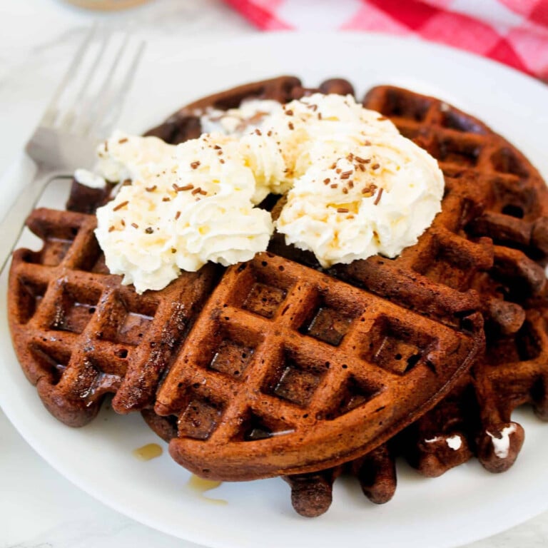 Chocolate waffle topped with whipped cream and chocolate sprinkles on a white plate.