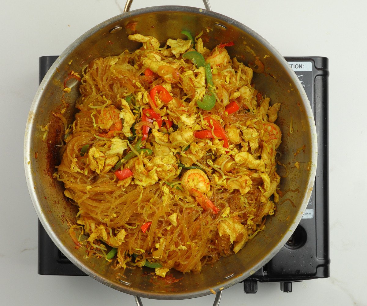 Singapore noodles, sauce, vegetables, chicken and shrimp cooking in a silver wok.