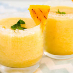 Two pineapple drinks in a glass cups with sugar on the rim, a pineapple chunk and mint leave.