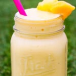 Mango smoothie in a glass jar with a pink straw and fruit in the rim on grass.