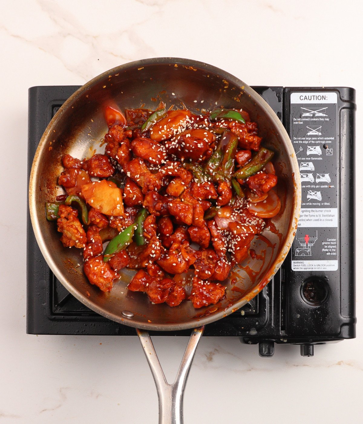 Red, green capsicum peppers, pineapples, crispy chicken, sesame seeds and sweet and sour sauce in a pan.