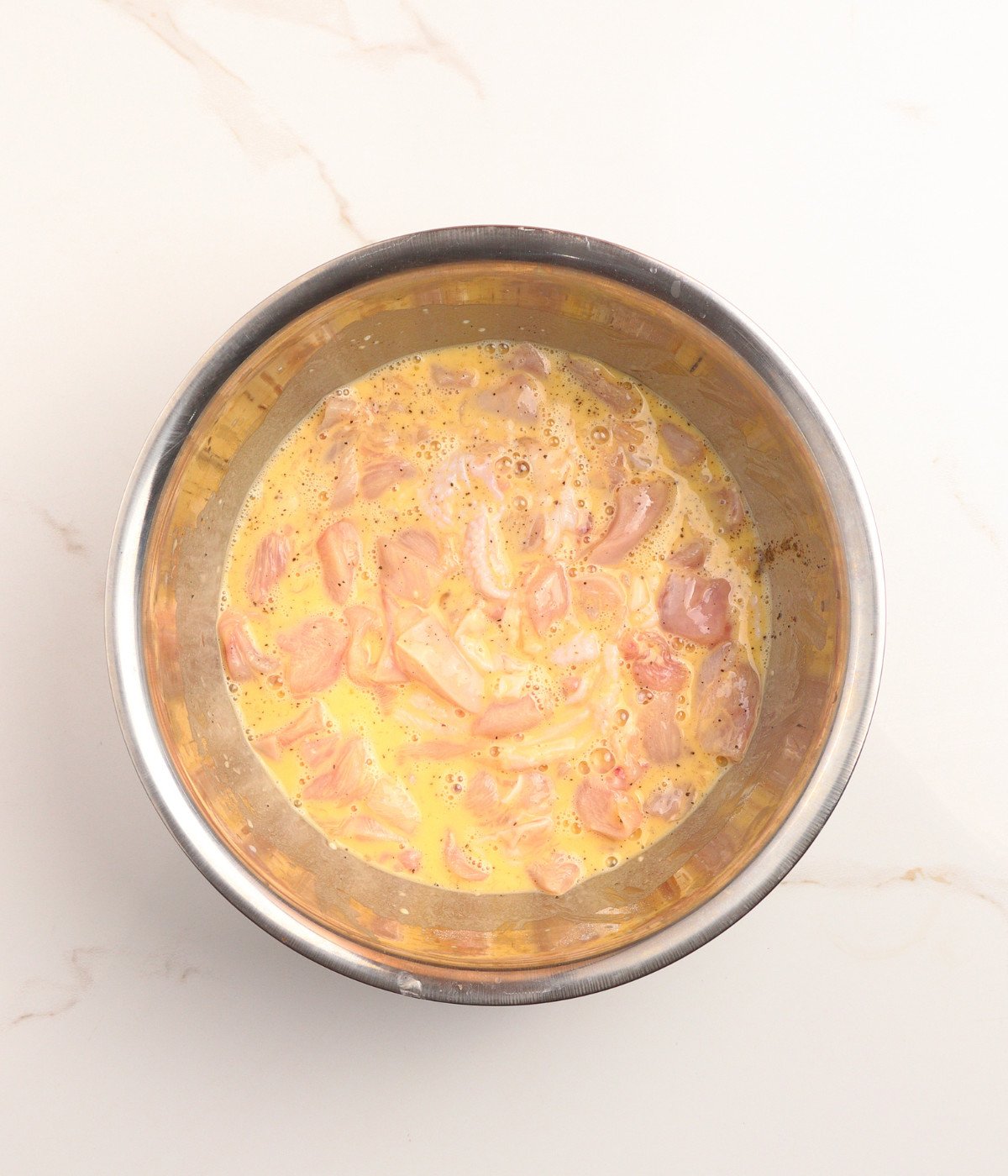 Uncooked chicken pieces, eggs, cornflour, water and seasoning in a silver bowl.
