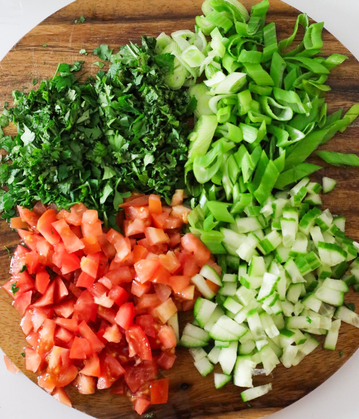 Fresh chopped herbs, scallions, tomatoes, and cucumbers on a brown chopping board.