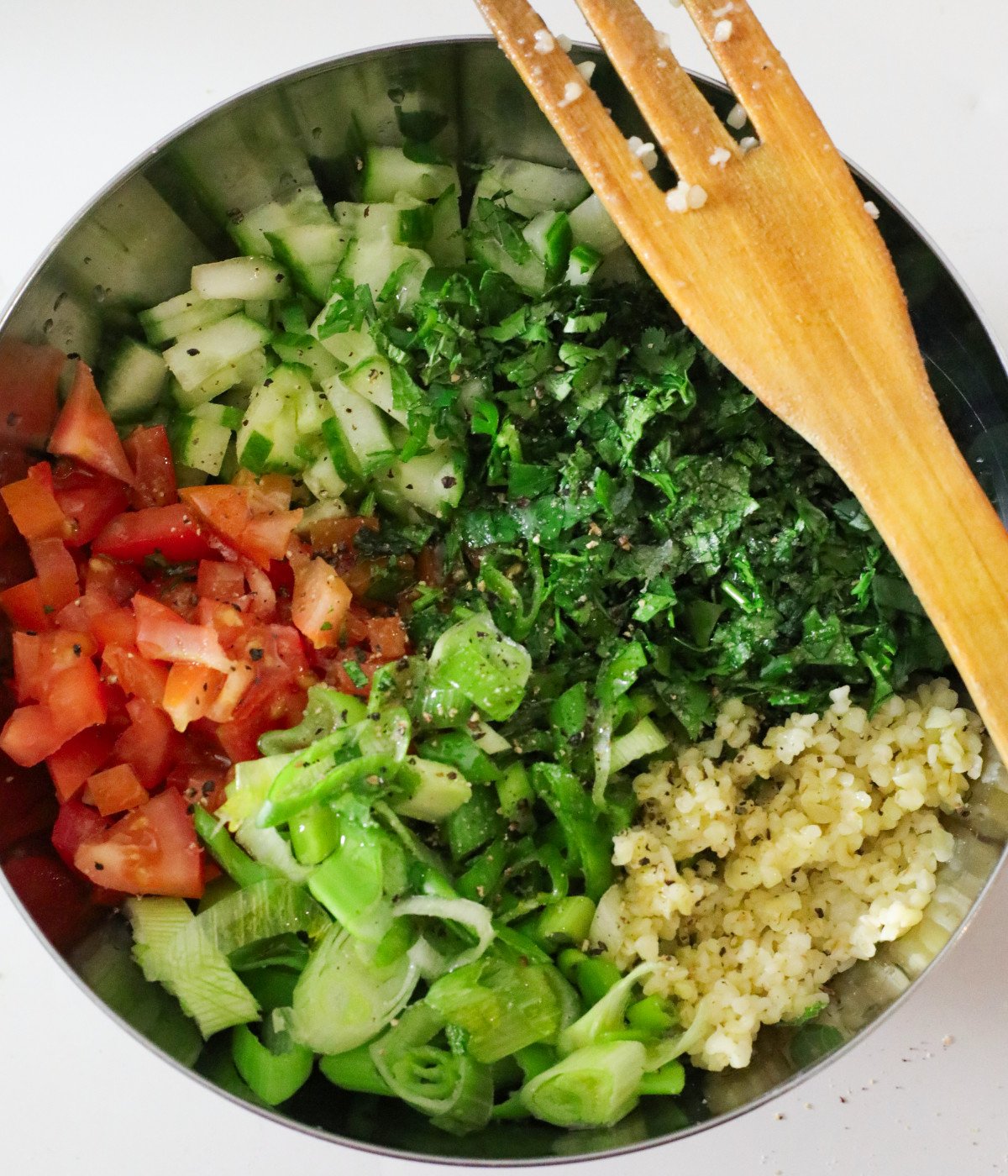Fresh chopped herbs, scallions, tomatoes, bulgur wheat dressing and cucumbers in a bowl with a mixing fork.