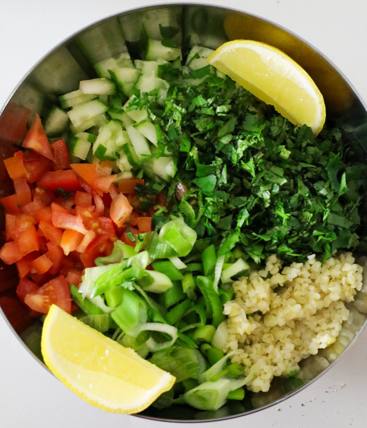 Fresh chopped herbs, scallions, tomatoes, bulgur wheat, lemon and cucumbers in a bowl.