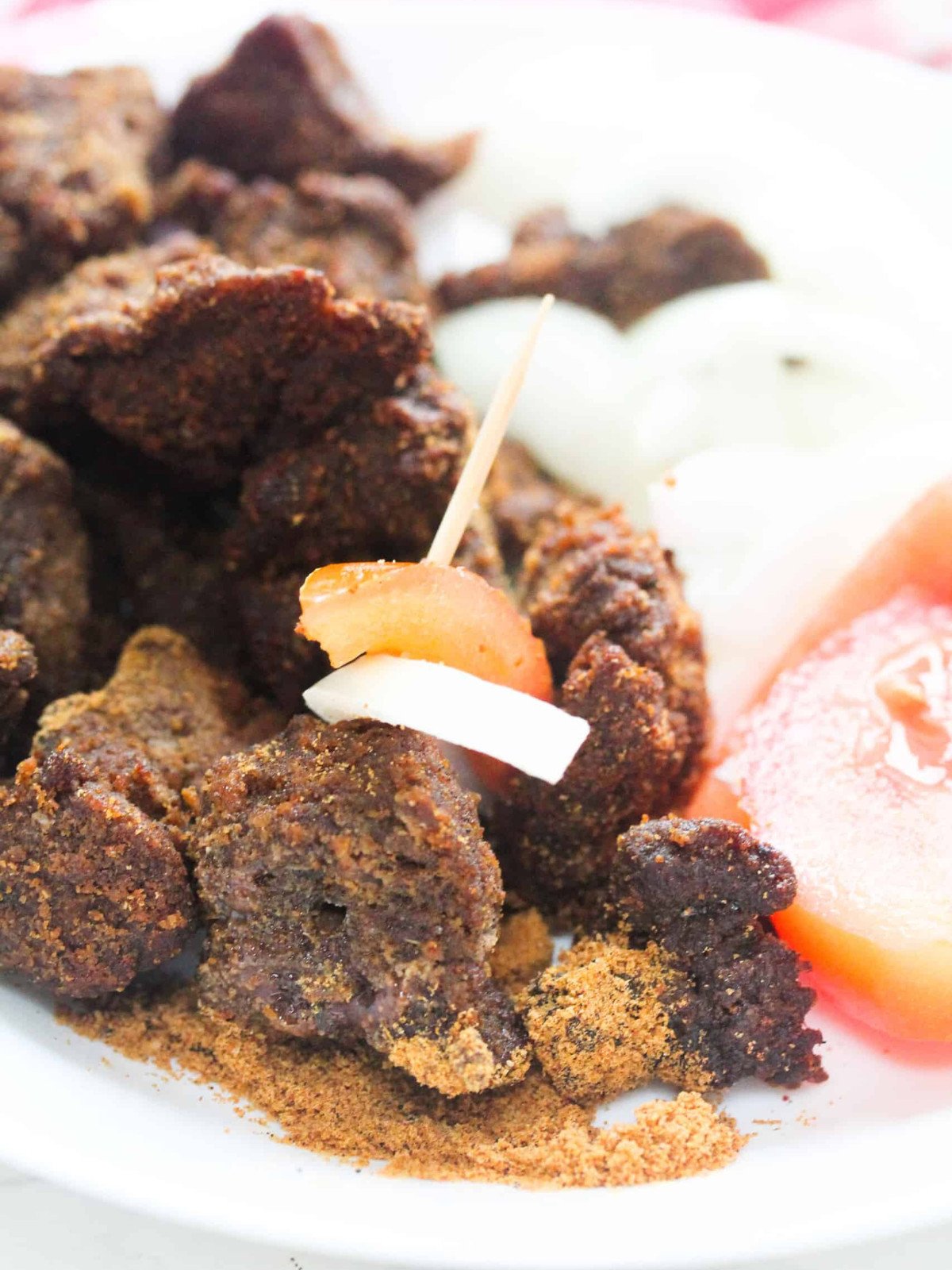 Suya beefs on a stick with omions and tomatoes on a white plate.
