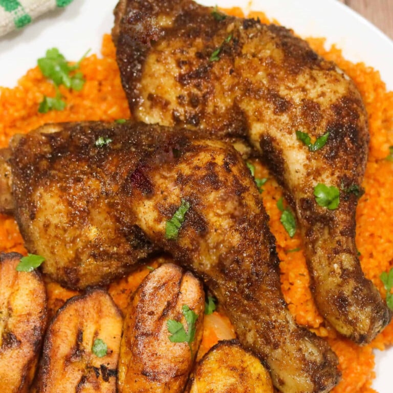 Suya chicken legs served with jollof rice, plantain and fresh parsley on a white plate.