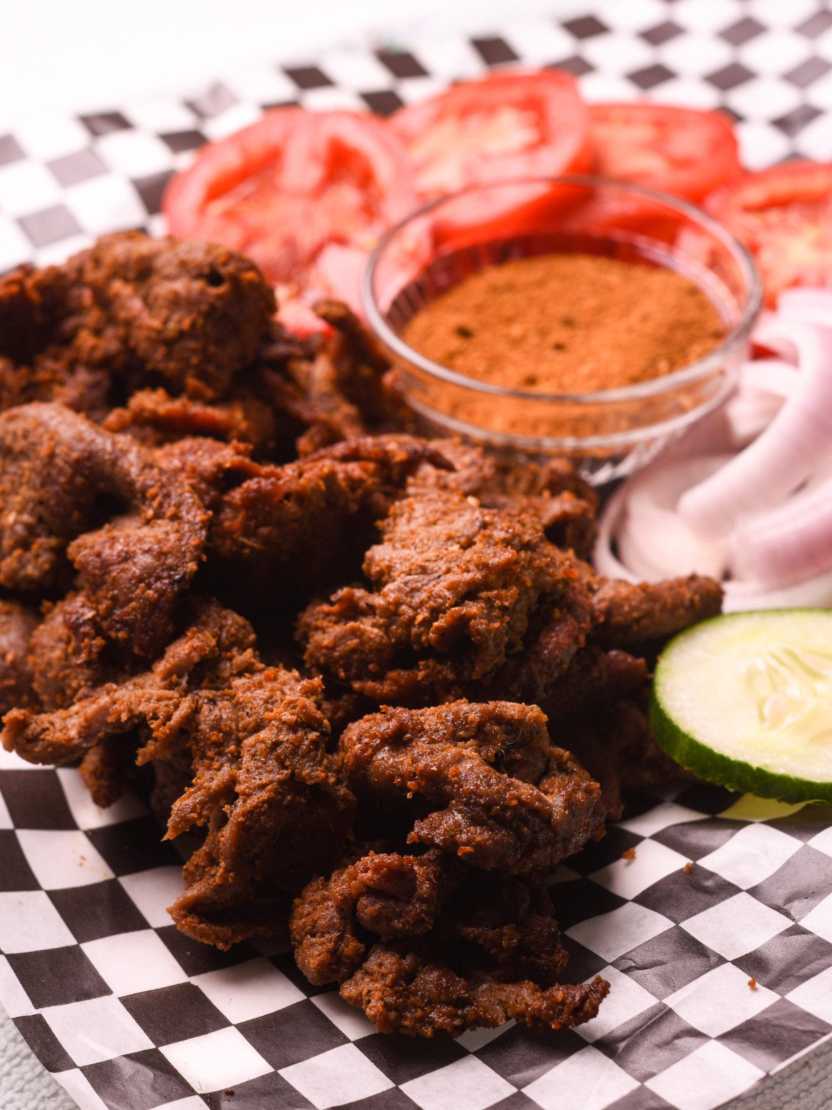 Suya meat, spice, cucumber, tomatoes and red onions on paper.