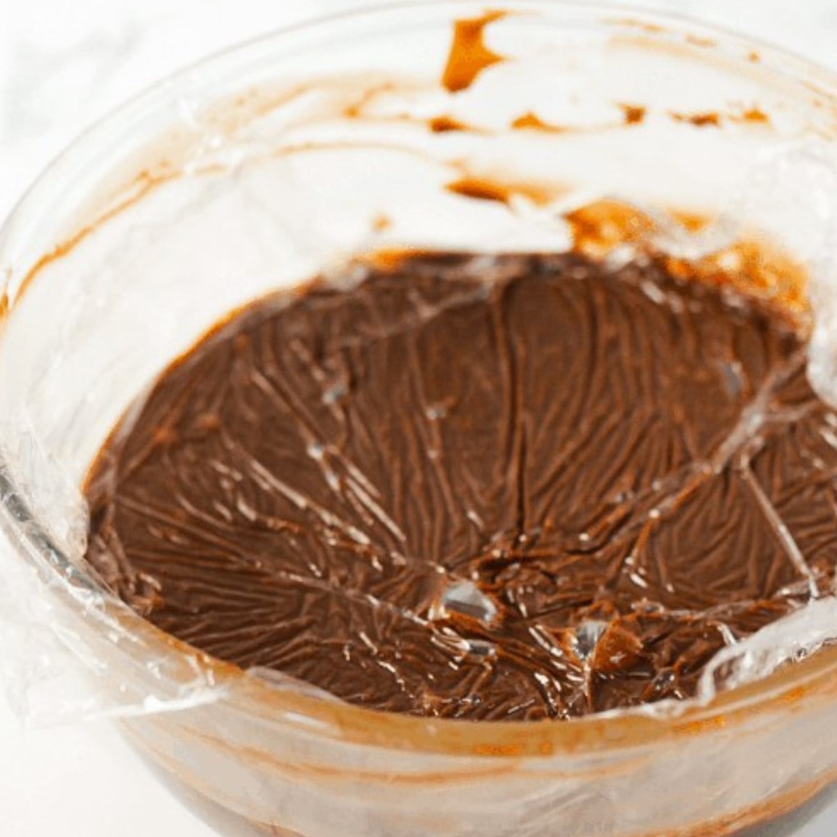 Hardened milk chocolate and heavy cream covered with plastic wrap in a glass bowl.