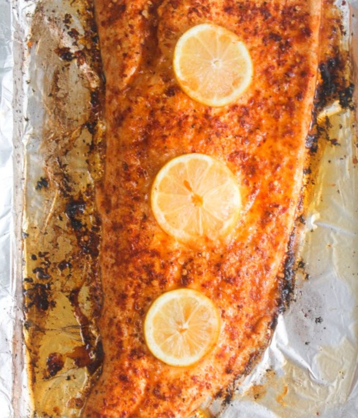 Half baked Salmon fillet on a baking tray lined with foil with three lemon slices on top.