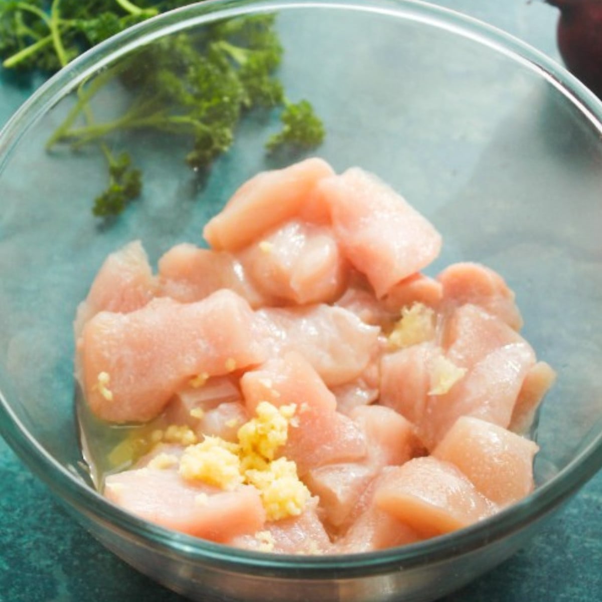 Raw chicken chopped in cubes with oil and garlic in a glass bowl.