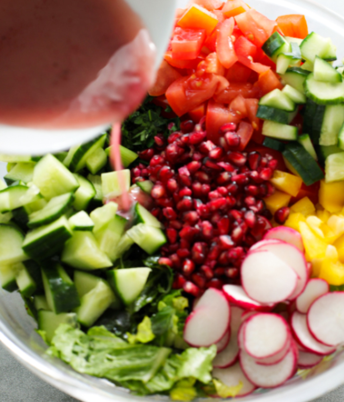 Tomatoes, cucumber, pomegranates, lettuce, radishes and yellow peppers and dressing in a glass bowl.