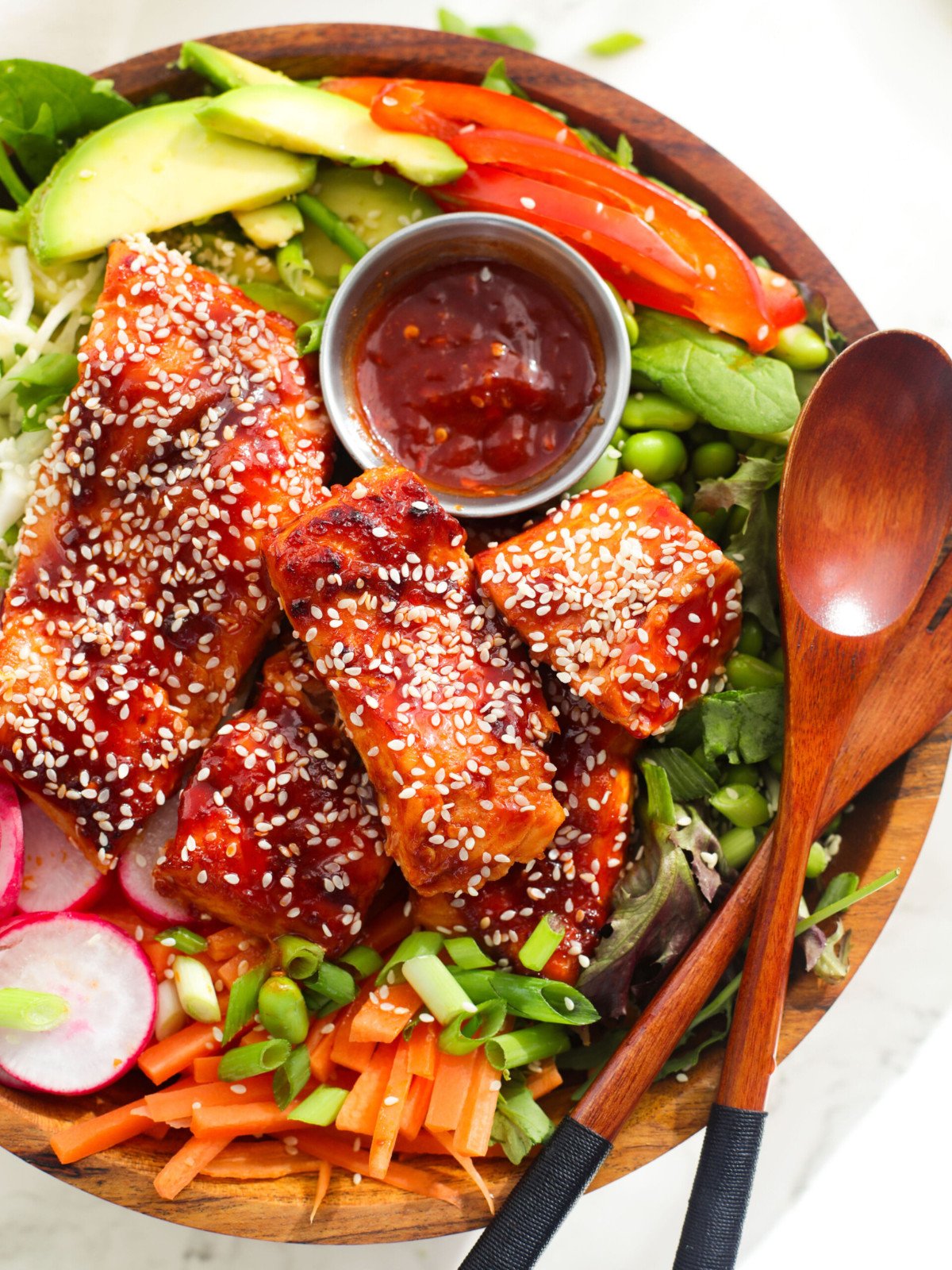 Firecracker salmon, avocado, edamame, tomatoes, carrots and sirracha dip in a bowl.
