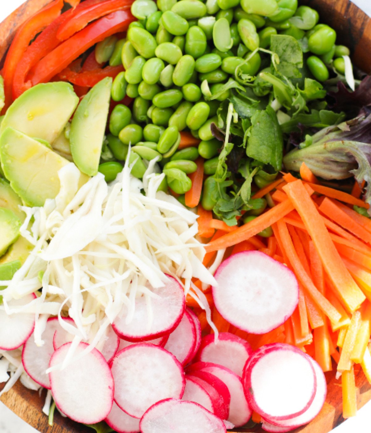 Red peppers, edamame beans, radish, avocado, carrots and cabbage in a bowl.