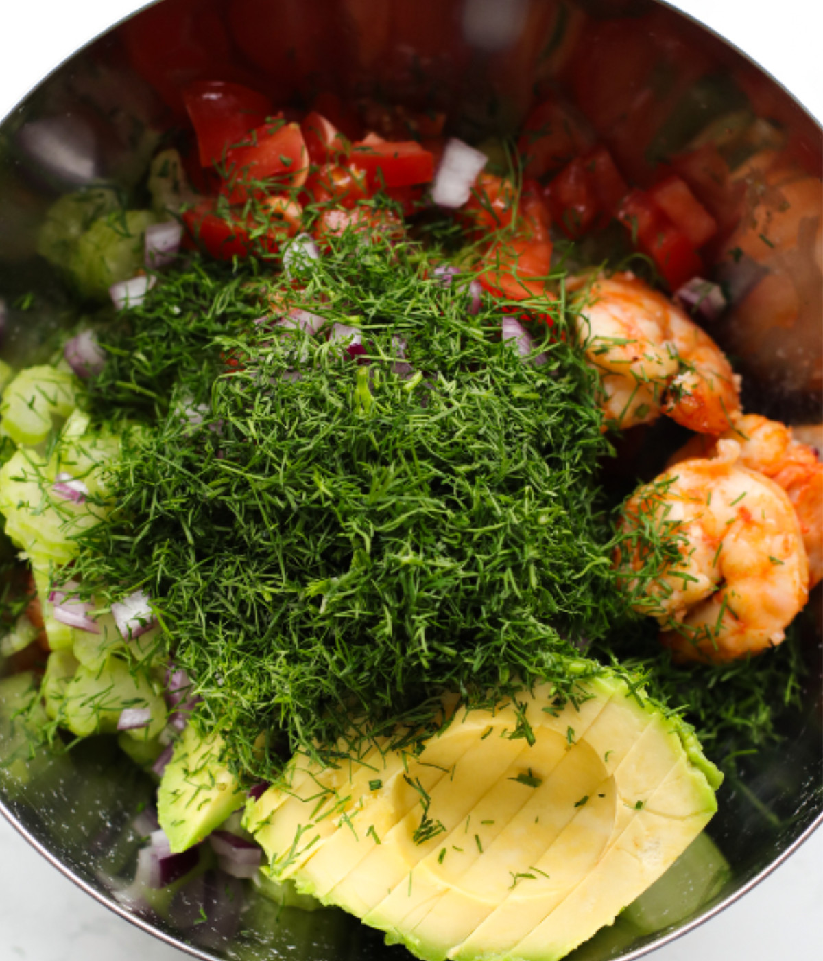 Cooked shrimp, dill, avocado, red onions, tomatoes and celery in a bowl.