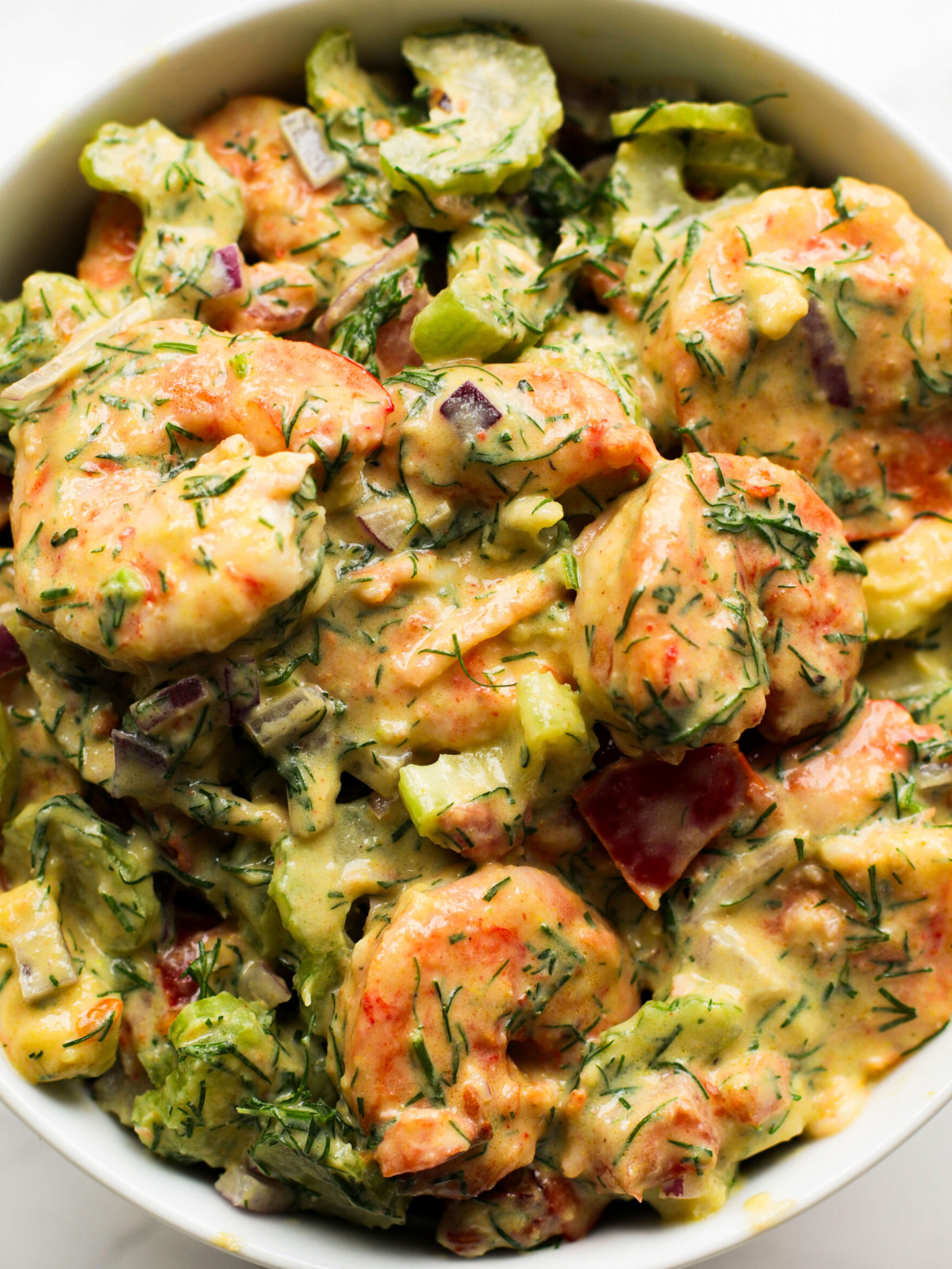 Large shrimp, avocado, celery, tomatoes, red onions and dill in a white bowl.