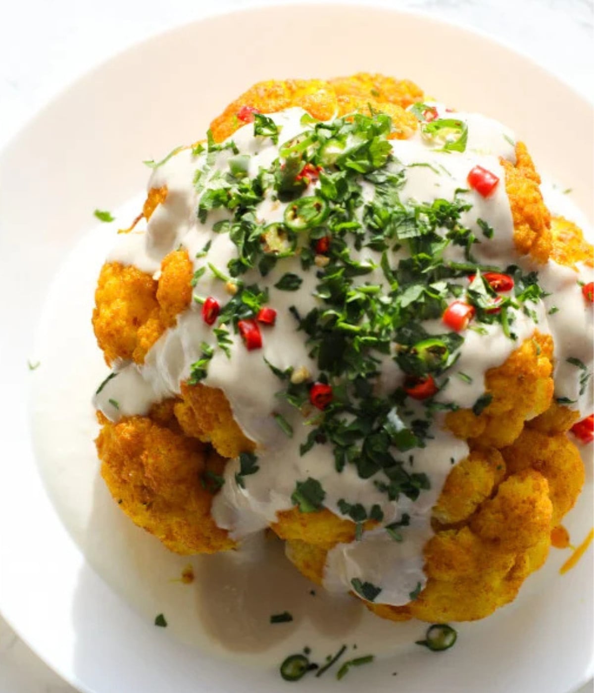 Baked turmeric cauliflower on a white plate with yogurt dressing underneath and tahini, herbs and chili on top.