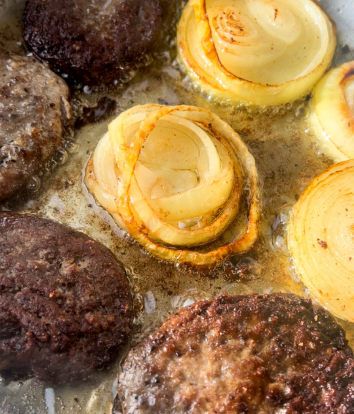 Beef patties and onion slices frying in a silver pan.