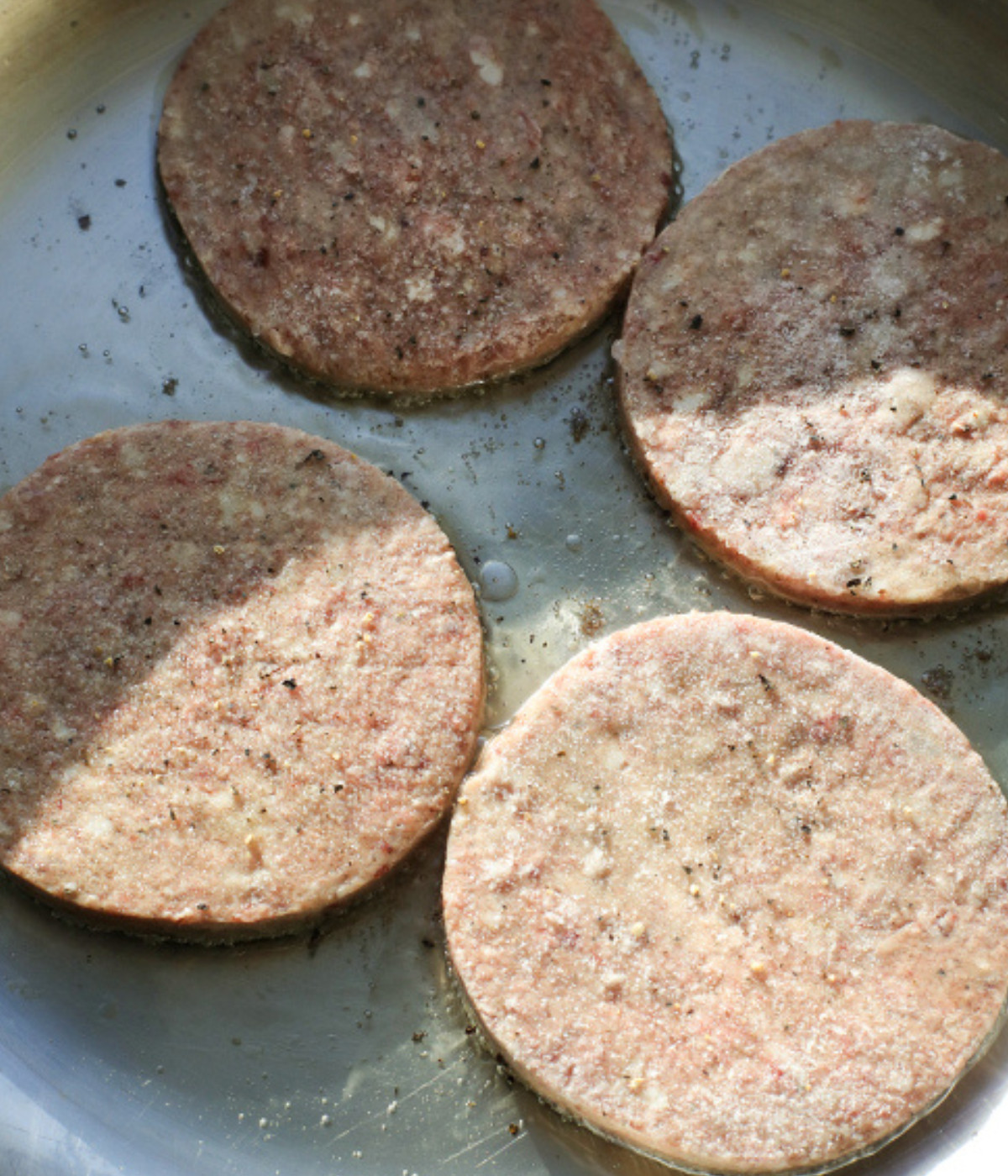 Beef patties frying in a silver pan with salt and pepper.