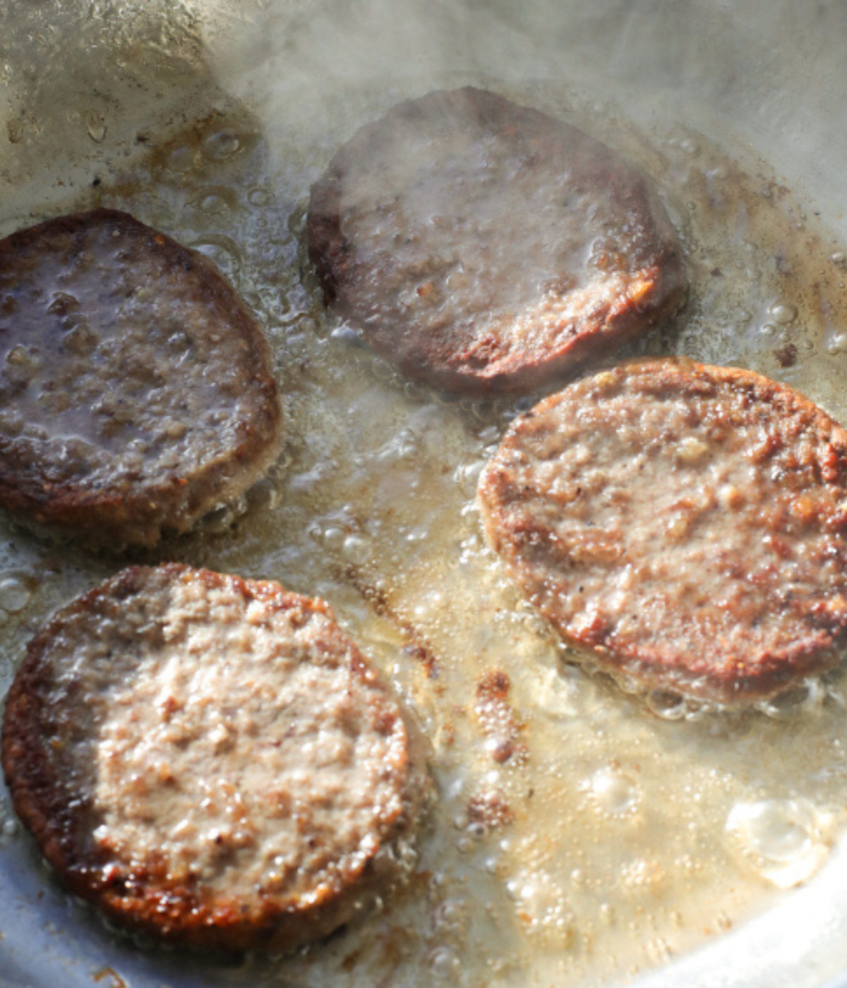 Beef patties frying in a silver pan.