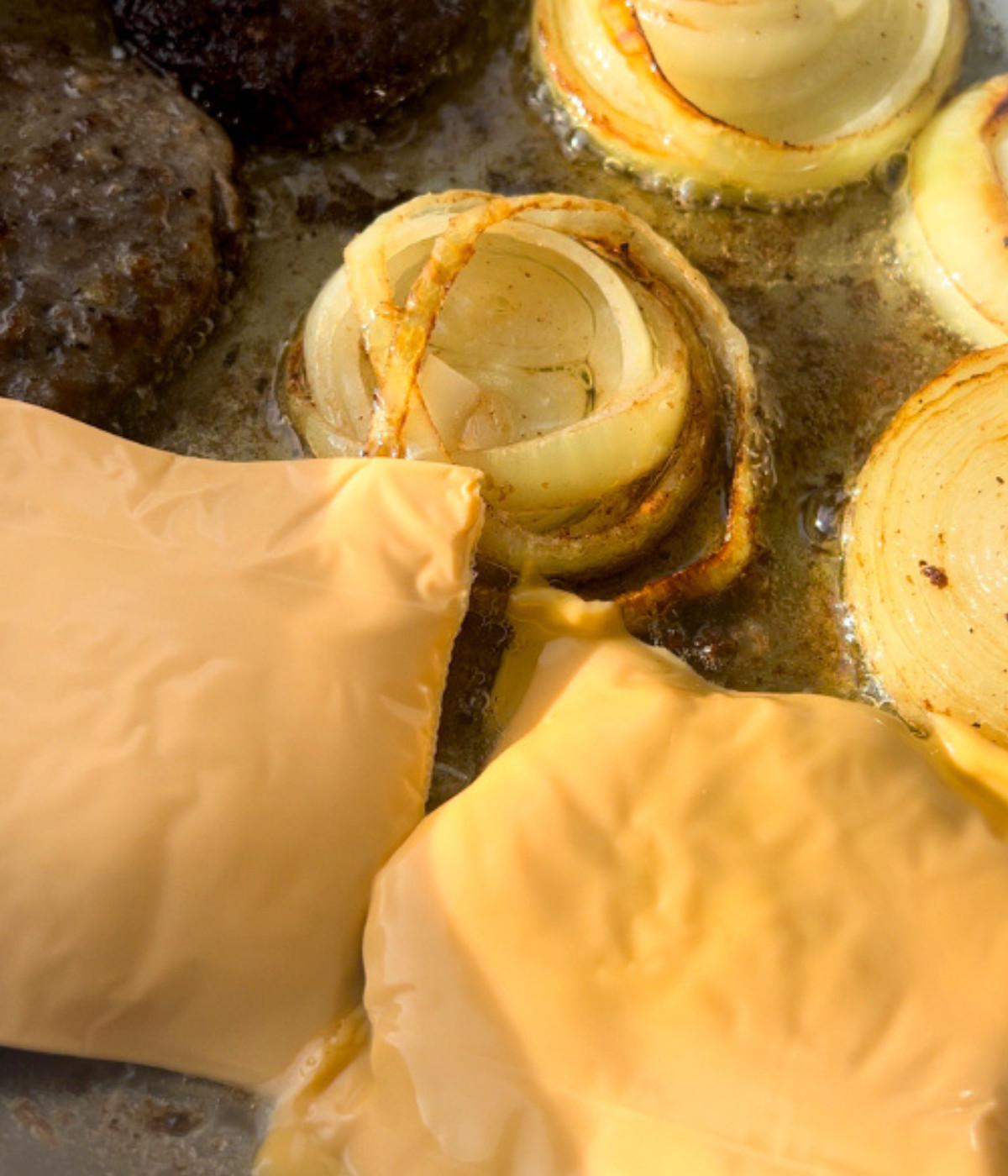 Beef patties with cheese slices on top and onions frying in a silver pan.