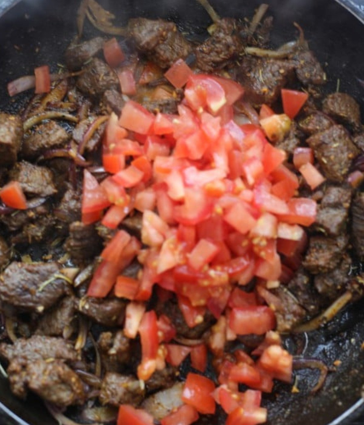 Chopped beef cooking in a black pan with chopped tomatoes.