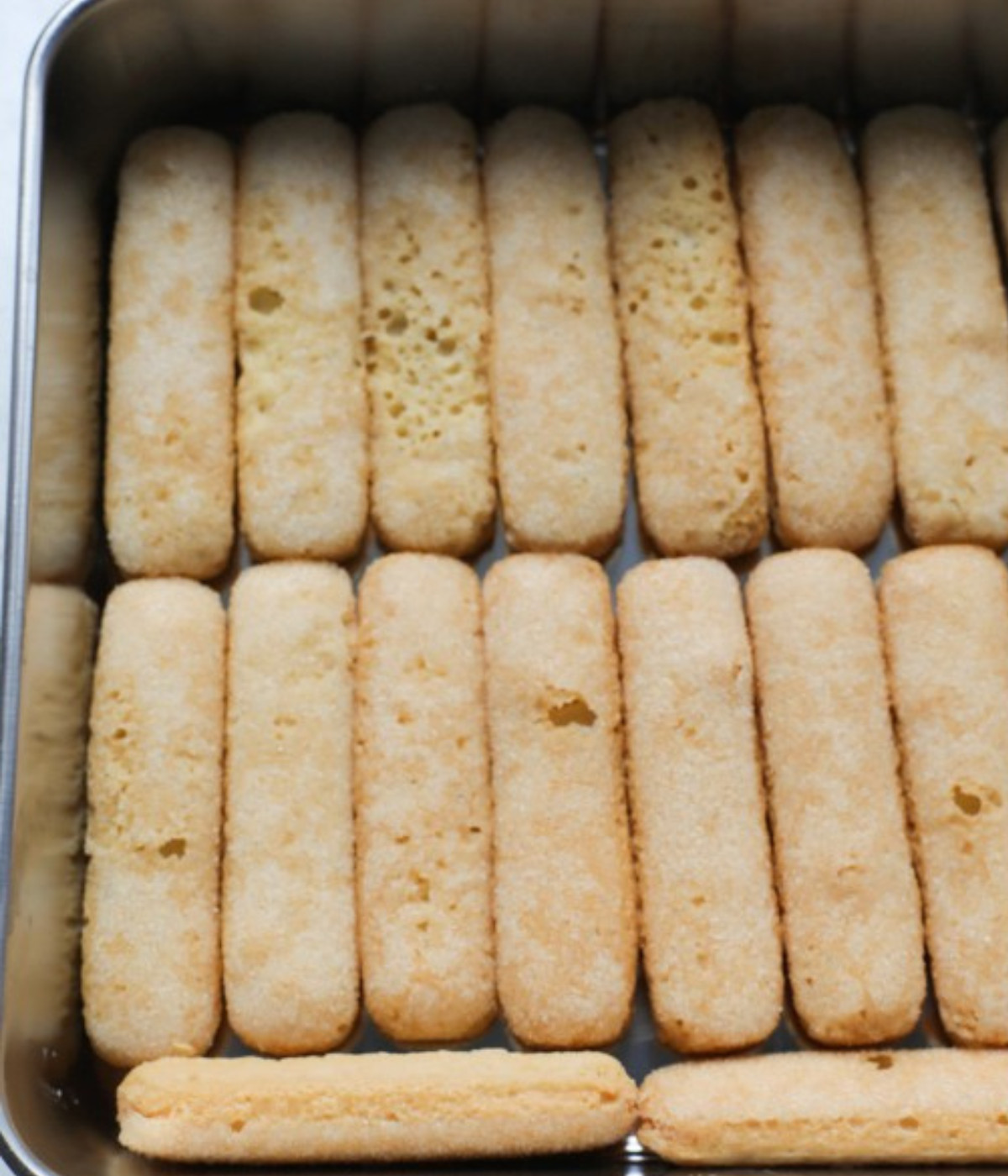 Soaked lady fingers laid in a square silver baking tin.